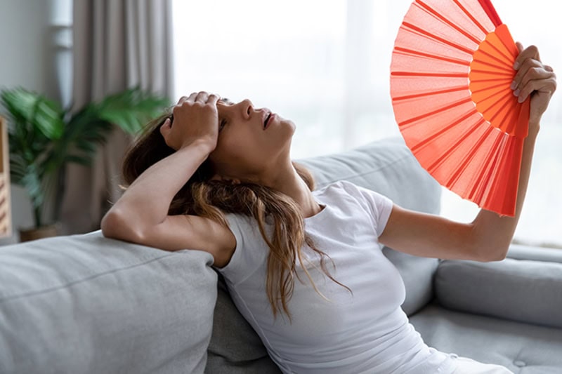 woman sitting on her couch looking exhausted as she fans herself with a paper folding fan.