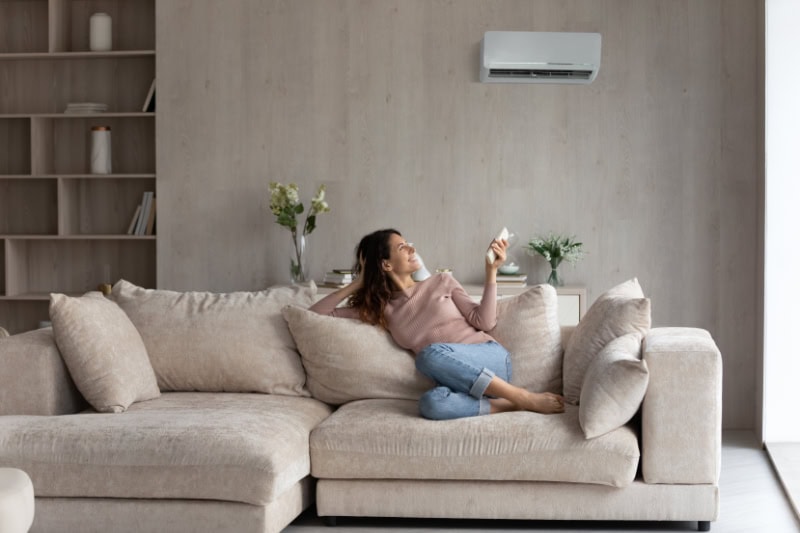 Relaxed young female homeowner sitting on huge comfortable couch, turning on air conditioner with remote controller, switching on cooler system, setting comfortable temperature in living room.